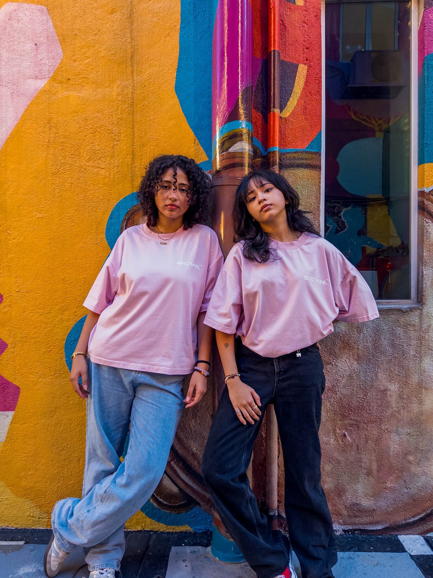 Both colourways worn side by side against a weathered wall, powder from the front and ink from the back with the full wordmark